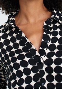 Camicia button-up bianca e nera con un motivo di grandi pois neri irregolari su uno sfondo beige chiaro.