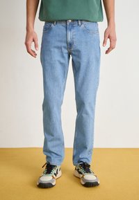 Jeans de denim azul claro com corte reto, cinco bolsos e detalhes de costura discretos. Usados com ténis multicoloridos e uma blusa verde.