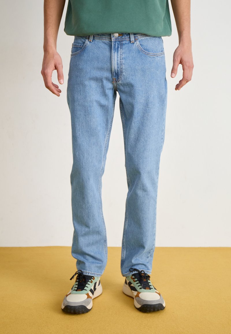 Jeans de denim azul claro com corte reto, cinco bolsos e detalhes de costura discretos. Usados com ténis multicoloridos e uma blusa verde.