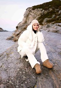 Biely páperový kabát s kapucňou, skombinovaný so smotanovými nohavicami a béžovými čižmami. Modelka má tiež na sebe béžovú pletenú čiapku a šál s třásňami.