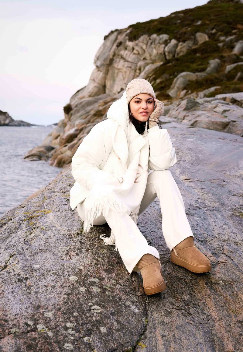 Biely páperový kabát s kapucňou, skombinovaný so smotanovými nohavicami a béžovými čižmami. Modelka má tiež na sebe béžovú pletenú čiapku a šál s třásňami.
