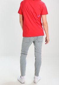 Camiseta de algodón roja con cuello redondo, que presenta un pequeño logo azul en la parte posterior; combinada con jeans ajustados de color gris claro y zapatillas blancas.