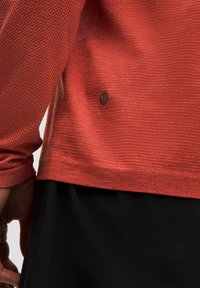 Top sportiv roșu cu mâneci lungi, cu un model texturat, guler rotund și un mic logo negru lângă tiv, asortat cu un articol vestimentar inferior negru.