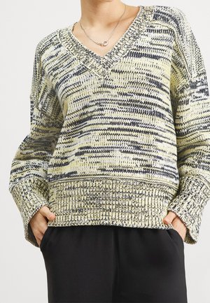 Person wearing a V-neck knit sweater with yellow and black mixed patterns and black pants, hands tucked into pockets.