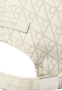 Μπεζ καπέλο Calvin Klein με υφή υφάσματος, διακριτικό επαναλαμβανόμενο μοτίβο λογοτύπου, ραμμένη ετικέτα бренда και ρυθμιζόμενο ασημένιο μεταλλικό κούμπωμα.