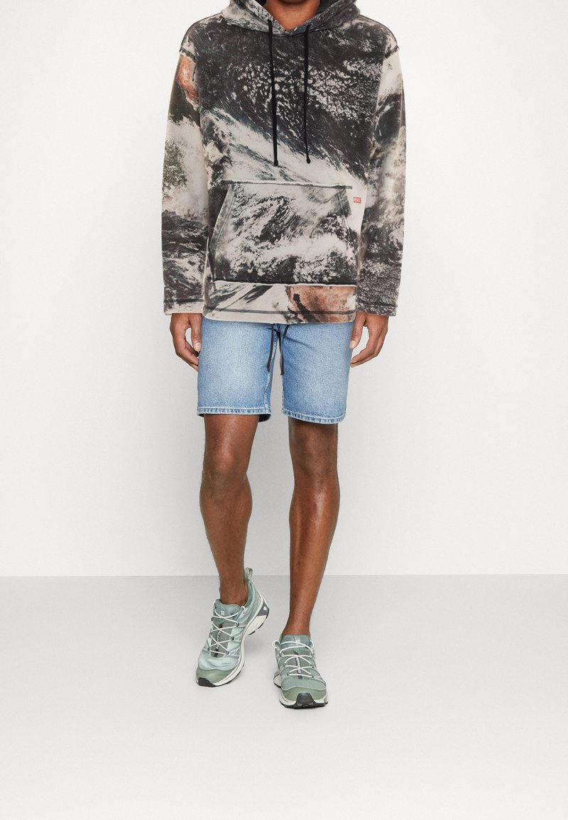 Persoon in een zwart-wit tie-dye hoodie, lichte blauwe denim shorts en lichtgroene sportschoenen, staand voor een plain witte achtergrond.