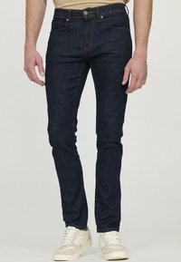 Jean en denim bleu foncé avec une coupe slim, featuring couture orange, design à cinq poches et fermeture à bouton à l'avant. Le modèle porte des baskets blanches.