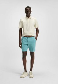 Paita polo dalam warna beige terang, dipadukan dengan celana pendek turquoise dan ikat pinggang cokelat. Model mengenakan sepatu putih, menampilkan penampilan yang santai dan modern.