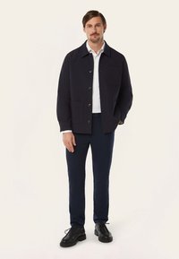 Marineblauwe jas met knopen aan de voorkant en een borstzak, gecombineerd met een witte blouse en marineblauwe broek, afgemaakt met zwarte schoenen.
