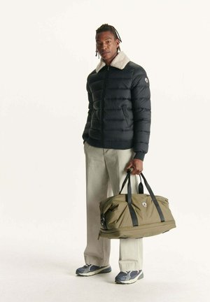 Homme portant une veste doudoune noire avec un col en fourrure blanche, un pantalon beige et des baskets, tenant un sac de sport vert olive avec des bretelles noires.