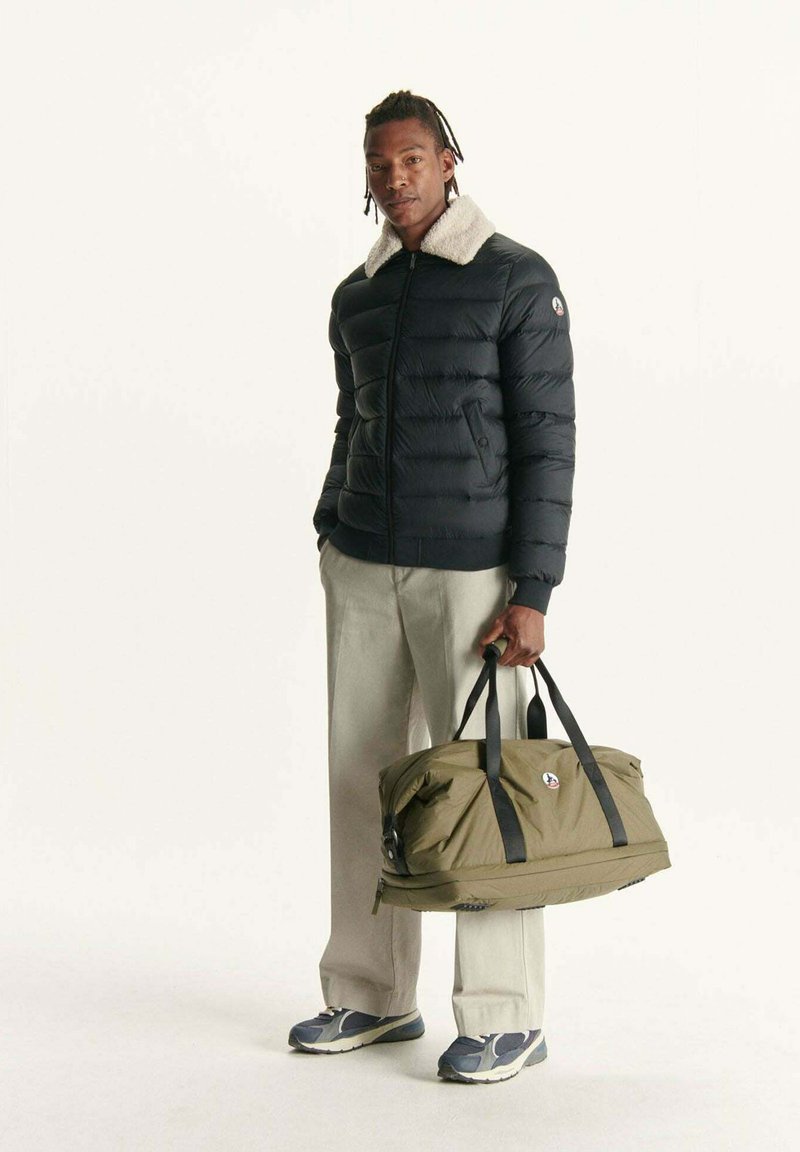 Uomo che indossa un giubbotto puffer nero con colletto in pelliccia bianca, pantaloni beige e sneakers, mentre tiene una borsa da viaggio verde oliva con cinghie nere.