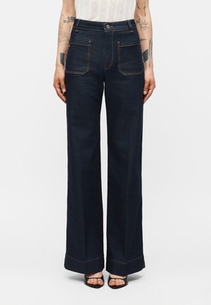 Žena má na sebe tmavomodré džínsy so širokými nohavicami s našitými vreckami vpredu a zatočenými lemami, v kombinácii s čiernymi otvorenými podpätkami.