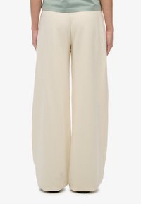 Brede beenbroek in een zachte beige stof. Heeft een hoge taille, een gladde textuur en een vloeiende silhoeet die tot de vloer reikt.