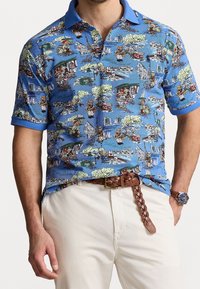 Lyhythihainen polo-paita sinisestä kankaasta, jossa on yksityiskohtainen, värikäs kuosi, joka esittää kohtia ja hahmoja. Yksivärinen sininen kaulus ja hihansuut.