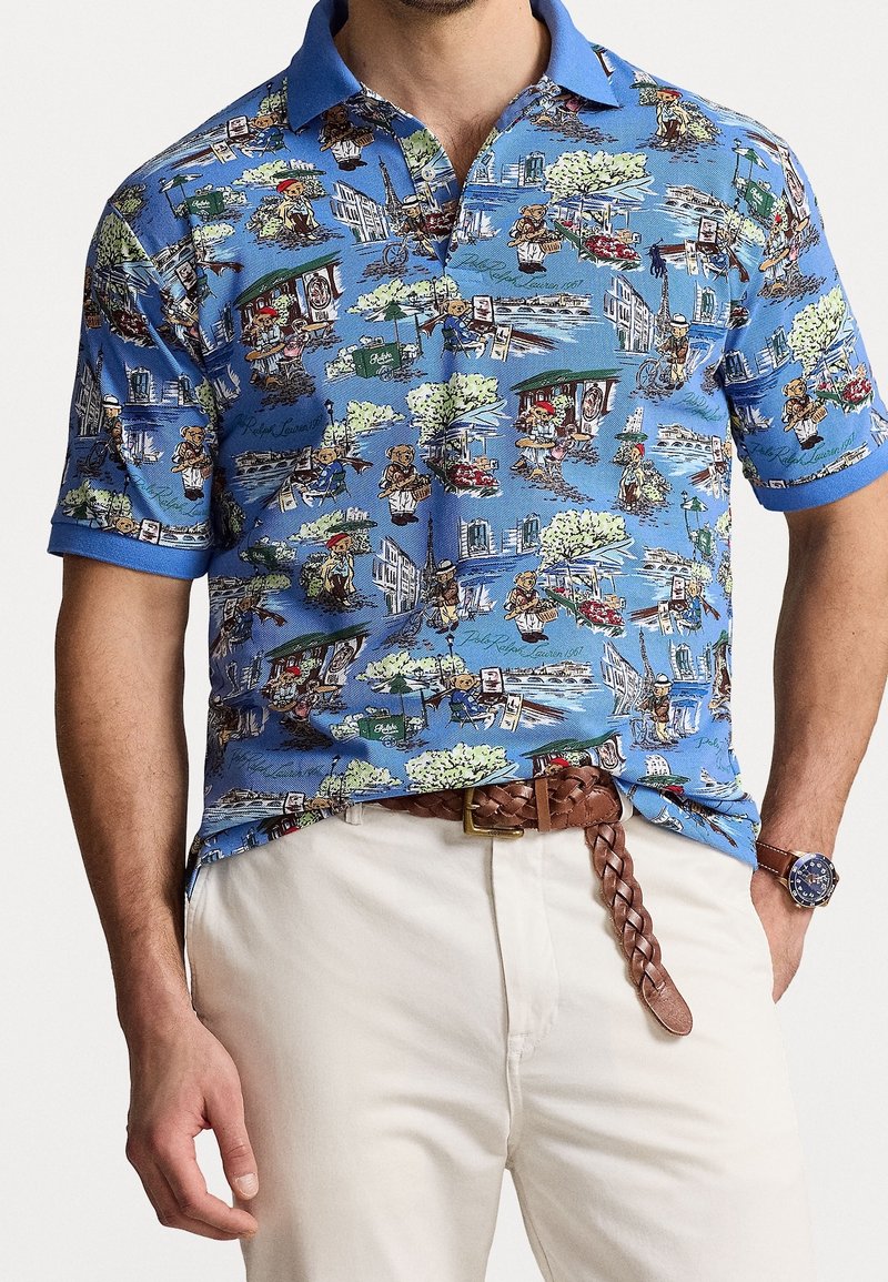 Lyhythihainen polo-paita sinisestä kankaasta, jossa on yksityiskohtainen, värikäs kuosi, joka esittää kohtia ja hahmoja. Yksivärinen sininen kaulus ja hihansuut.