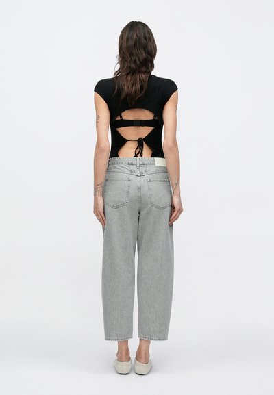 Haut noir côtelé avec dos découpé et détail noué, associé à un jean gris à coupe ample et des chaussures de couleur claire. Fond neutre.