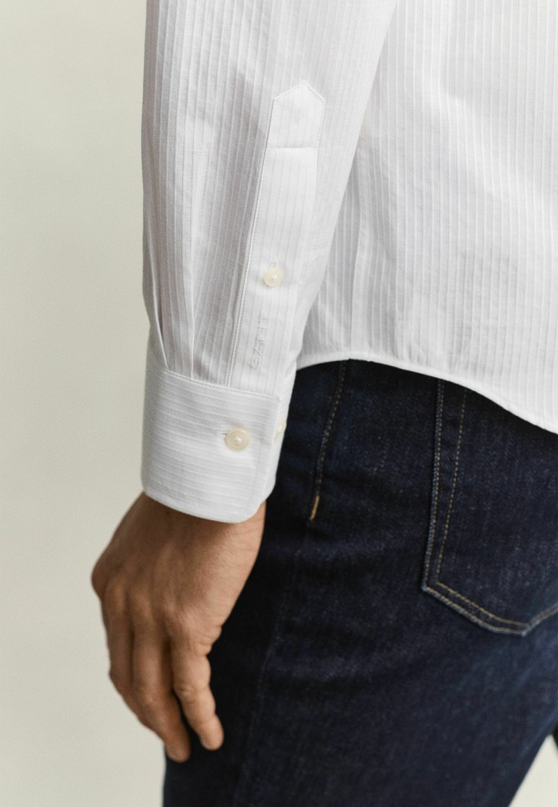 Man's left hand beside hip wearing white striped dress shirt with buttoned cuff and dark blue denim jeans.