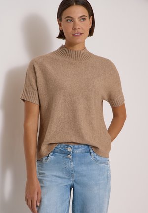 Femme portant un pull en tricot beige à manches courtes et un jean taille haute bleu clair, debout devant un fond uni.