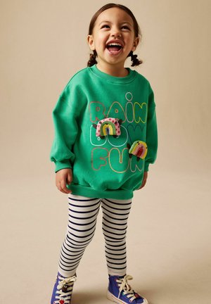 Kleines Kind lacht, trägt einen grünen Pullover mit bunten Regenbögen und dem Schriftzug "RAINBOW FUN", gestreifte Leggings und blaue Turnschuhe.