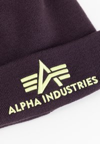 Cappello beanie di colore viola scuro realizzato in tessuto lavorato a maglia, con un logo ricamato di colore verde chiaro e la scritta "ALPHA INDUSTRIES" sul risvolto.