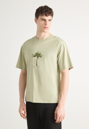 T-shirt en coton vert clair à manches courtes, avec un graphique de palmier et du texte en dessous en marron foncé, coupe décontractée, texture lisse.