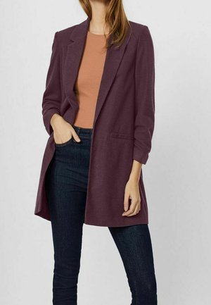 Mujer que lleva un blazer largo color ciruela sobre una blusa color óxido y vaqueros ajustados azul oscuro, con una mano en el bolsillo y la otra relajada.