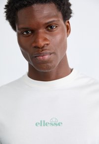 Tricou alb din bumbac, cu un logo "ellesse" de culoare verde deschis în centru. Guler rotund, textură netedă, design ajustat.