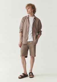 Cămașă ușoară din in maro peste un tricou alb, asociată cu pantaloni scurți bej și sandale maronii. Design simplu cu detalii cu nasturi și mâneci rulote.