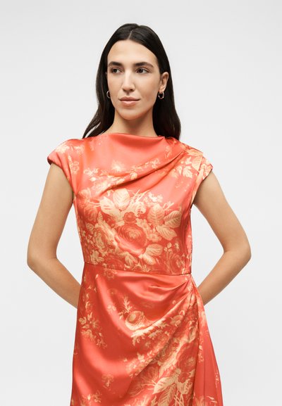 Γυναίκα με μακριά σκούρα μαλλιά που φοράει κοραλλί σατινέ φόρεμα χωρίς μανίκια με λουλουδάτο μοτίβο, στέκεται με τα χέρια πίσω από την πλάτη της, κοιτάζοντας αριστερά.