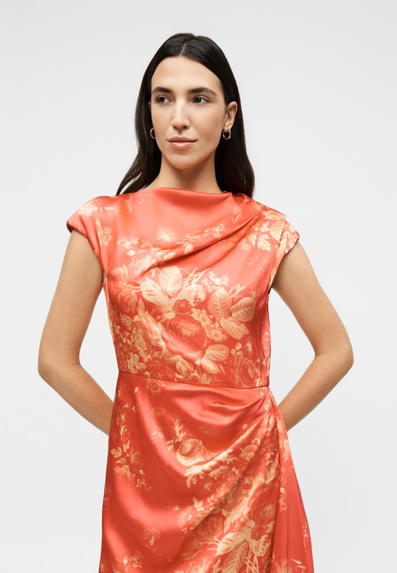 Woman with long dark hair wearing sleeveless coral satin dress with floral pattern, standing with hands behind back, looking left.