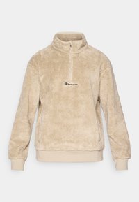 Béžový fleecový pulóver s krkom na zips, ktorý má mäkkú, štruktúrovanú tkaninu a malú vyšitú logo na hrudníku.