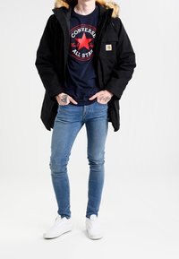 Parka negra con capucha forrada de piel, cierres de cremallera y botones a presión, llevada sobre una camiseta oscura con un logo de estrella. Jeans azules ajustados y zapatillas blancas.