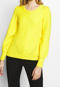 Gelber Strickpullover mit Rundhalsausschnitt, gerippten Bündchen und strukturiertem Muster. Mit seitlichen Mesh-Details und taillierter Silhouette.