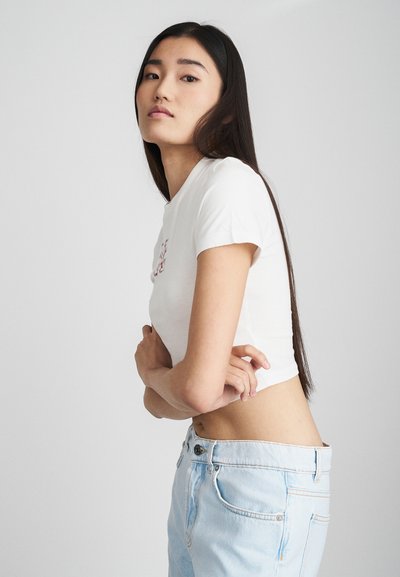 T-shirt blanc court à manches courtes, avec un petit graphique rouge. Le modèle porte un jean en denim taille haute de couleur bleu clair.