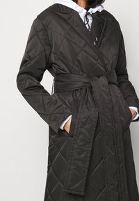 Černý prošívaný dlouhý kabát s páskem uvázaným v pase, nošený p�řes bílou mikinu s černými šňůrkami, ruce v kapsách kabátu.