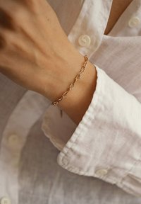 Pulsera de oro rosa con formas ovaladas entrelazadas y acentos de estallido solar, llevada en una muñeca sobre la manga de una camisa de lino blanca.