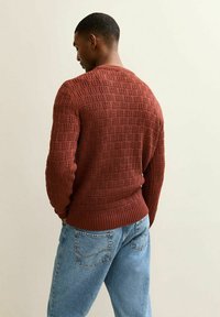 Gebreide roodbruin trui met een gestructureerd patroon en ribbelzoom, gecombineerd met losse lichtblauwe jeans. Zicht van achter.