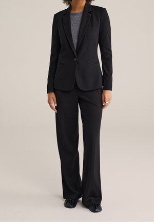 Czarny garnitur na miarę z marynarką zapinaną na jeden guzik i dwoma przednimi kieszeniami, zestawiony z prostymi spodniami. Szara koszula wewnętrzna o fakturze.