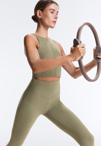 Ženska v zeleni vadbeni obleki drži pilates obroč z obema rokama in stoji v širokem razširjenem položaju vaje.