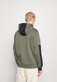 Sudadera verde oliva con capucha y mangas negras; cuenta con una textura suave y puños acanalados. La vista trasera muestra un diseño minimalista sin logotipos.