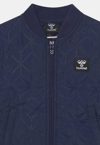 Marineblauwe gewatteerde jas met diagonale stiksels, ritssluiting aan de voorkant, geribde kraag en een zwarte patch met logo met een witte bij en hummel-tekst op de borst.