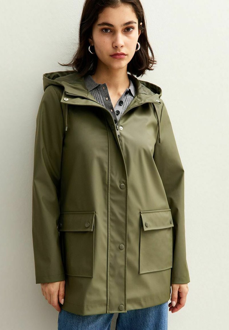 Chaqueta impermeable de color verde oliva con capucha, bolsillos frontales y cierres de botón. Presenta una textura suave y un diseño de longitud media.