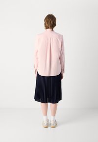 Camicia button-up rosa chiaro con orlo curvo, abbinata a una gonna plissettata blu navy. Calzini bianchi e scarpe chiare completano il look.