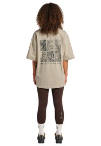 Personne aux cheveux bouclés portant un t-shirt beige oversize avec six panneaux de cartes en noir et blanc dans le dos, des leggings marron, des chaussettes blanches et des baskets noires.
