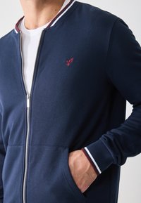 Mornarska zip-up dukserica s ovratnikom i manžetama u bijelim i burgundy prugama. Sadrži mali crveni izvezeni logo i bočne džepove.