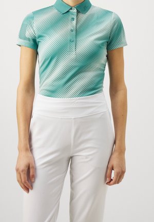 Vrouw die een aangesloten teal poloshirt draagt met diagonale witte strepen en witte broek met hoge taille, staand tegen een effen achtergrond.