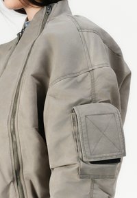 Giacca bomber grigia con chiusura a zip, texture imbottita e una grande tasca con velcro sulla manica.