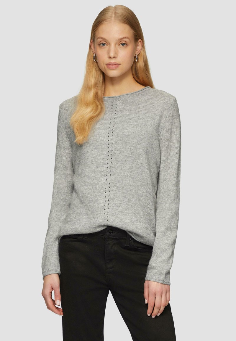 Pull en tricot gris clair à manches longues, col rond, avec une ligne verticale de petits trous décoratifs au centre. Associé à un pantalon noir.