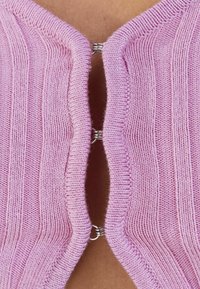 Top de punto rosa claro con rayas verticales, que presenta una abertura en la parte delantera y cierres a presión metálicos para el ajuste. Textura suave.