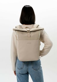 Beige rol-top rugzak gemaakt van duurzaam materiaal, met verstelbare schouderbanden en een voorvak; gladde textuur met een minimalistisch ontwerp.